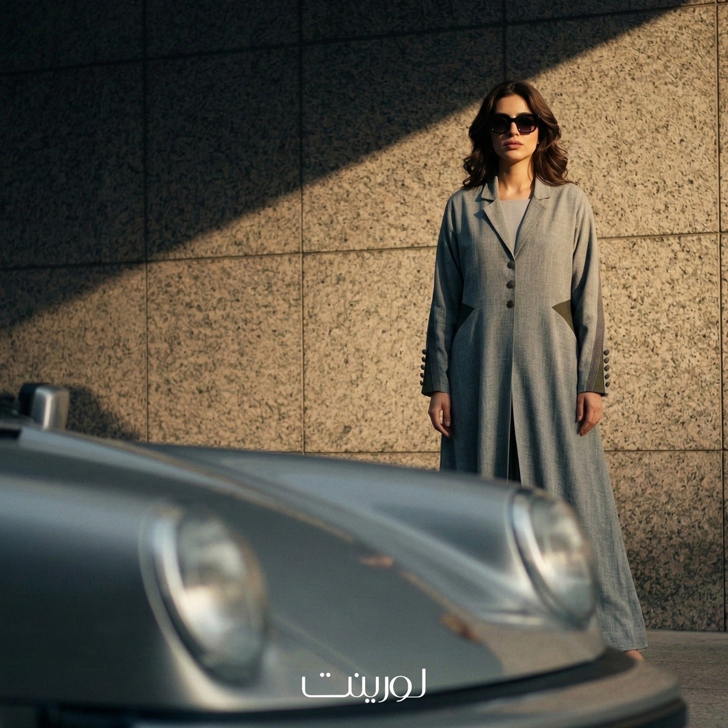 Woman in a long coat standing next to a car with a textured wall background